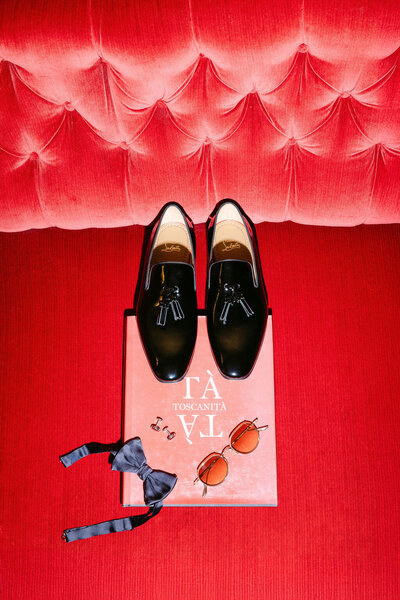 Groom's Christian Louboutin shoes on a red sofa, along with his sun glasses and bowtie.
