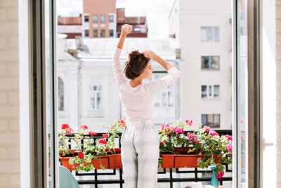 Woman stretching on a balcony, facing away, surrounded by blooming flowers in planter boxes. She's wearing a white top and plaid pants, with buildings visible in the background on a sunny day.