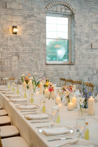 a wedding reception table is set with white plates, glassware, and small favor bottles, decorated with pastel floral arrangements, an indoor wedding reception at grbic events in st. louis