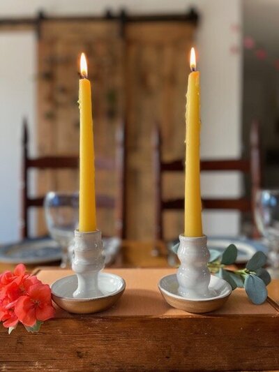 a set of two beeswax candles in white pottery, handmade candlestick holders.  Set on a table with flowers around it.