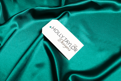 Holly Taylor Designs logo the name written elegantly and a needle with thread displayed on a tag laying on green slik
