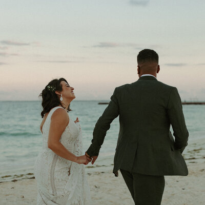 The bride and groom run hand in hand on the beach in Cancun, Mexico. The colors for the sunset were the perfect shade of pink and blue giving the most beautiful filmy vibe.