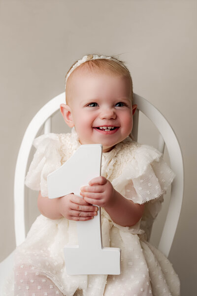 Happy toddler in tan hoodie outfit sitting inside wooden prop bowl against warm brown backdrop, captured in a classic cake smash portrait.