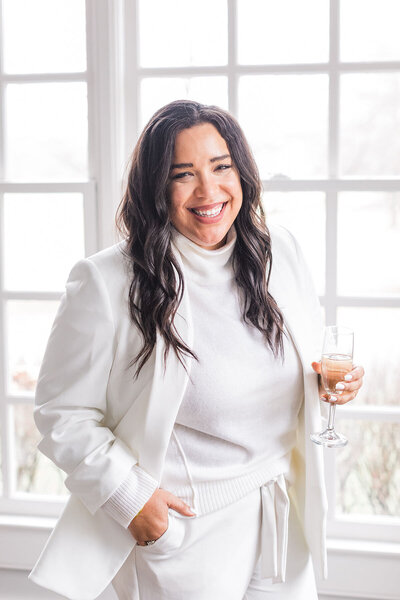 Natasha Churches, owner of Aisle & Co., poses for a headshot in all white while holding champagne.