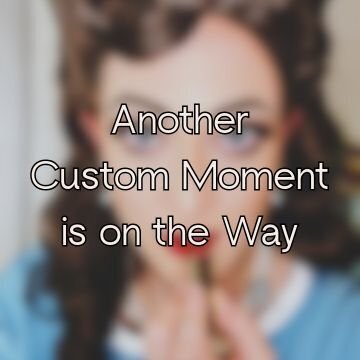 Blurry image of a brunette vintage-style bridal wig with lipstick being applied, featuring centered text overlay reading 'Another Custom Moment is on the Way' — placeholder for future wig gallery additions.