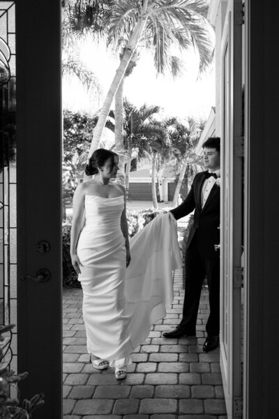 groom holds brides dress as she walks through doorway