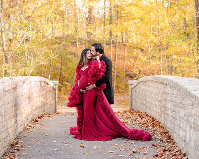 Pregnang mother in a fluffy red dress posed with her husband at Inniswood. He is kissing her on the cheek.