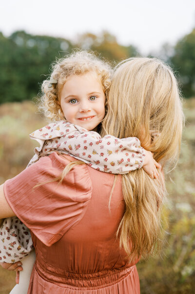 Little girl with curly hair and bright eyes hugs around Mom's neck during playful family session in Raleigh NC