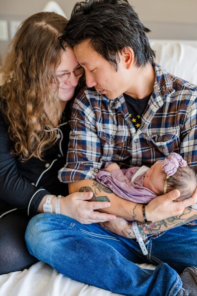 Mom and dad sitting next to each other on hospital bed holding newborn baby. 