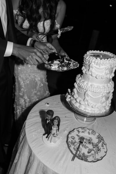 Up close of the bride and groom cutting their wedding cake, with bobble head cake topper on the table, taken on flash in black and white.