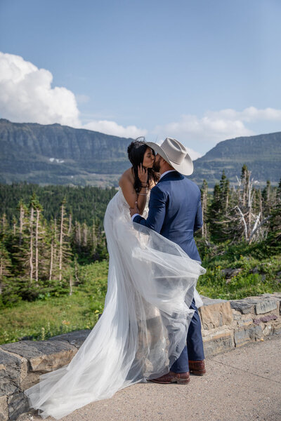 Bride and groom share a romantic kiss on a stone overlook in Glacier National Park, Montana, with rugged mountain peaks in the background. The bride’s sheer wedding dress flows in the wind, while the groom, dressed in a blue suit and cowboy hat, embraces her during their adventure elopement.