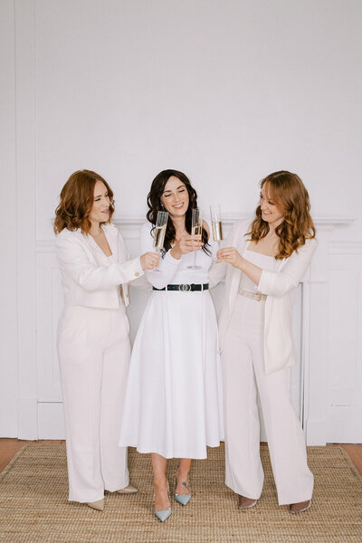 Three women in white attire stand together, holding champagne glasses for a toast, against a plain white background—perfect imagery for a wedding planner website.