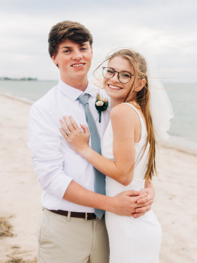 A young couple in wedding attire stands on a sandy beach, smiling and embracing. Featuring an Eau Claire Wedding Photography duo, the bride wears glasses and a veil, while the groom sports a light blue tie. The ocean shimmers in the background.