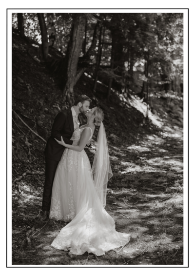 An elegant first look photo of bride and groom kissing underneath trees on a forest trail. captured in exquisite detail by a documentary photographer in New England. The groom's hand, adorned with a wedding ring, rests on the bride's lace gown as her veil drapes elegantly, revealing intricate embroidery and feather details.