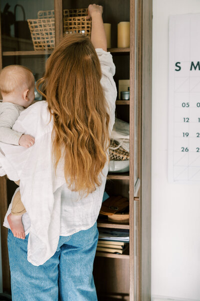 Woman with long wavy hair holding a baby while reaching up to a high shelf in a cabinet filled with baskets, candles, and household items - Family Autism Support Coach at Chantal Hewitt Coaching