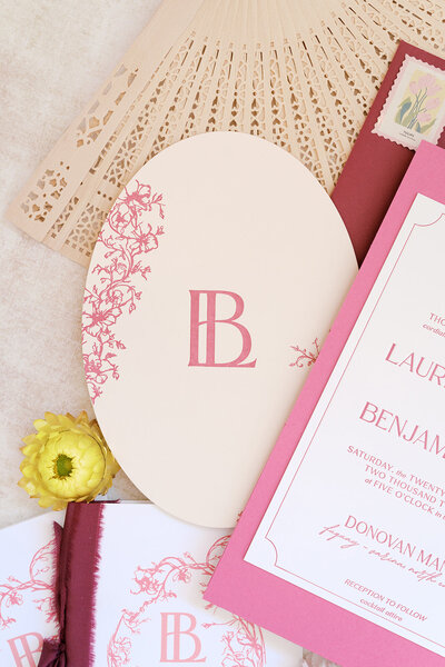 Custom monogram wedding invitation suite by Grey Cove Designs styled with a laser-cut fan, blush envelope, and floral crest illustration designed for a fall celebration.