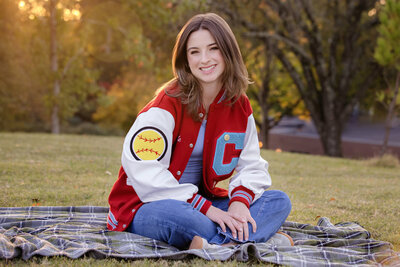 Confident HS senior in a softball Charlotte Catholic HS letterman jacket sitting on a blanket in green grass at UNCC smiling in soft autumn light.