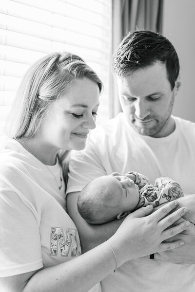 A couple stands in front of a window. The father holds their newborn baby while the mother wraps her arm around his arm. They both look at their baby with a soft smile in this tender shot captured by a photographer specializing in newborn photography in Austin, TX