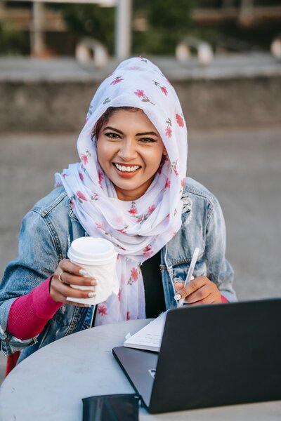A smiling woman is wearing a head scarf and holding a cup.