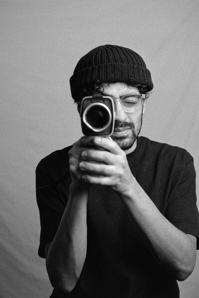 Black-and-white split portrait of two photographers with vintage cameras, wearing beanies and glasses. One holds a Canon film camera, the other a classic video camera, capturing timeless moments with an analog touch.