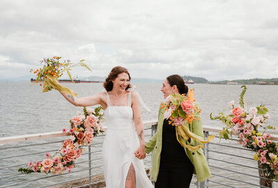 Two brides celebrating at the wedding altar. They are looking at each other with joyful expressions and raising their bouquets in the air. The bride on the left is wearing an all-white dress, and the bride on the right is wearing a light green suit jacket with a black shirt and black pants. The couple is flanked by two large ceremony flower installations that are built onto the railing of the wooden dock that the couple is standing on. The flowers are a colorful blend of pink, peach, green, purple, and orange flowers. In the background is a gorgeous view of the Columbia River Gorge in Astoria, Oregon. On the water is a large cargo ship. In the distance are some hills.