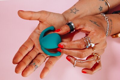 The camera looks down on two tattooed hands wearing several rings, with bright red nails. A teal vibrator is centered in the palm of the right, open hand.