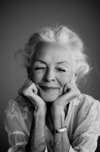 An elderly woman with closed eyes and a peaceful smile, resting her chin on her hands. She wears a light-patterned blouse in this black-and-white photo by Sweet Pea Ink Creative.