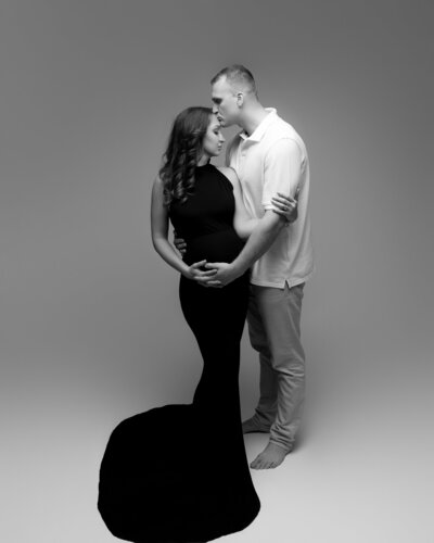 woman wearing sleeveless, mermaid style maternity gown embracing with her husband in white during studio maternity photoshoot