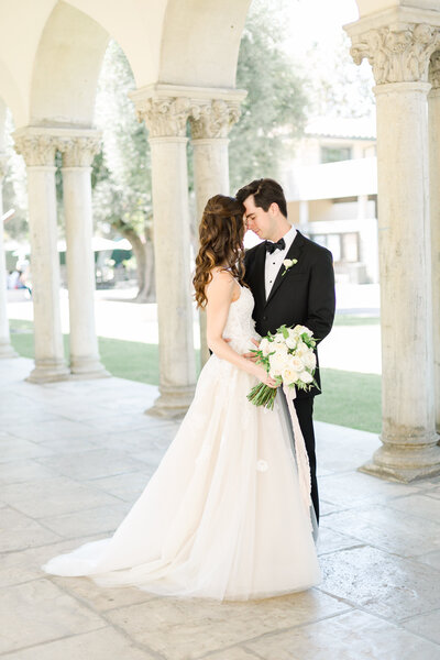 Bride and groom stand forehead to forehead while holding wedding bouquet under arched walkway