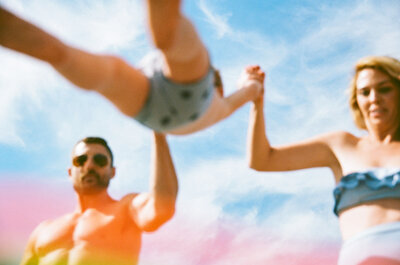 out of focus photo of mom and dad in swimsuits swinging their toddler over the camera