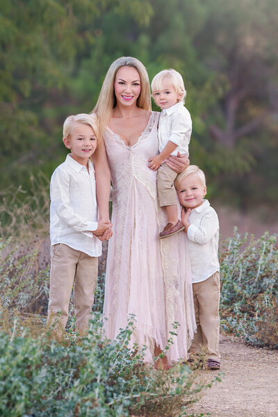 A mother with blonde hair wearing an off-white lace dress standing in a field with her three young sons all in white linen button up shirts and khaki pants while smiling