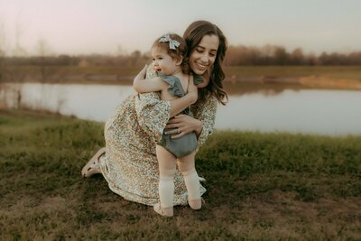 A woman in a floral dress kneels on grass by a lake, smiling as she hugs a toddler in a bow and romper. Captured beautifully by a Warner Robins family photographer.