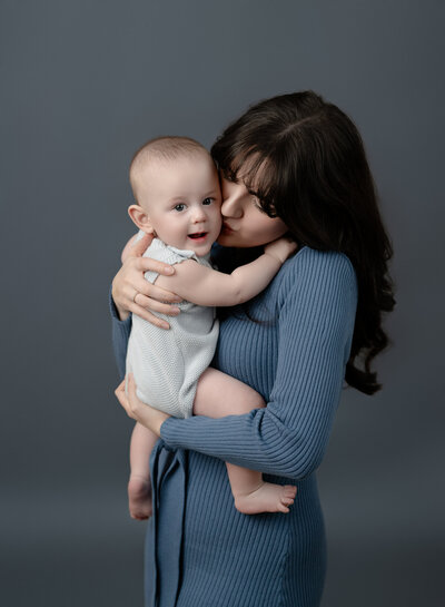 Mommy and me photo with Mom kissing baby boy's cheek while hugging him closely. Studio photo taken by Jenni Nikki Photography.