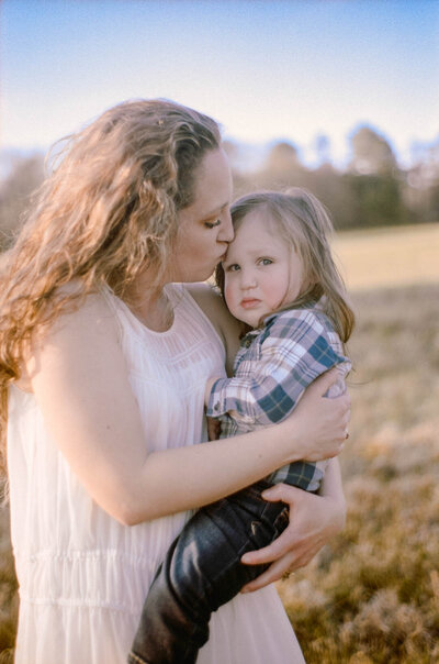mother kissing her son on the forehead while family session takes place in a field