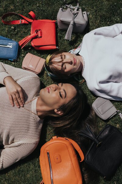 Two women lay with eyes closed on grass surrounded by vibrant hand bags