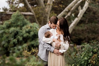 A family moment with a man, woman, and baby embracing outdoors during a newborn lifestyle photography session.