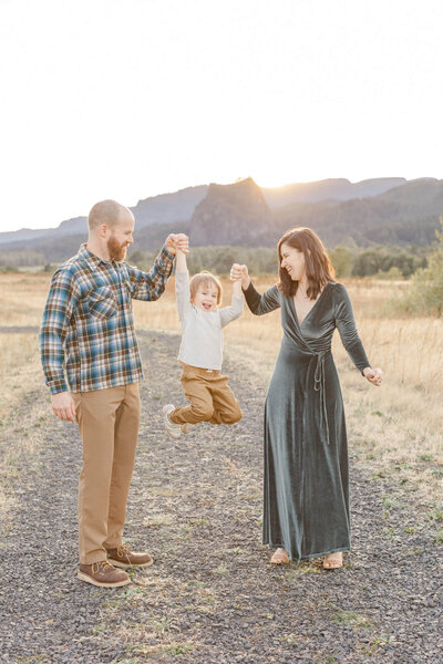 Mom and Dad holding toddler's hands in between them and raising him up at their fall family photo session in the Columbia River Gorge at sunset. 