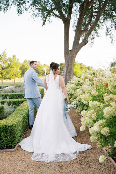 Bride and groom walking hand in hand along a hydrangea-lined garden path during their East Coast–inspired wedding.
