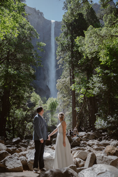 A couple in wedding clothes looks back over their shoulders at a waterfall