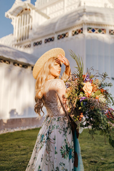 Bride holding a giant bouquet of flowers and wearing a wide-brimmed hat standing in the sun