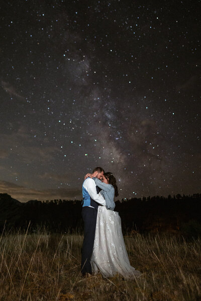 A bride and groom embrace under a sky filled with stars and the Milky Way during their Montana elopement, captured by Sydney Breann Photography in a glowing nighttime portrait.