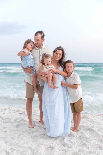 Happy family of five standing on the beach at sunset, mom holding daughter, dad holding toddler, son smiling by the ocean, soft pastel outfits and ocean backdrop.