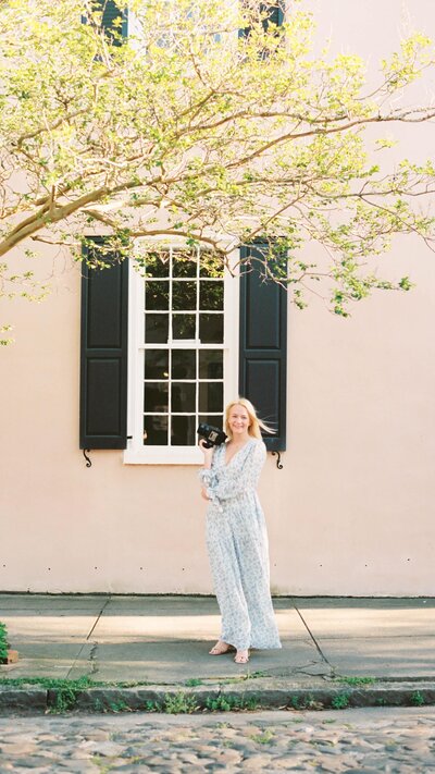 Rhode Island Wedding Photographer | Deirdre in a light dress stands smiling on a cobblestone street. A pink wall with a black shuttered window and tree branches frame the scene