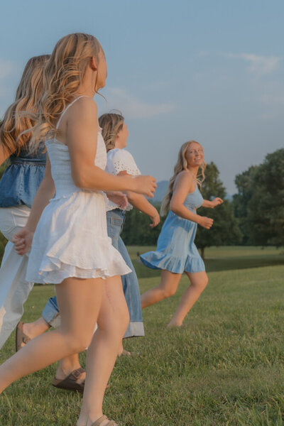 Four best friends run in a field together with trees in the background. They look at each other and smile while they run in Johnson City, TN. 