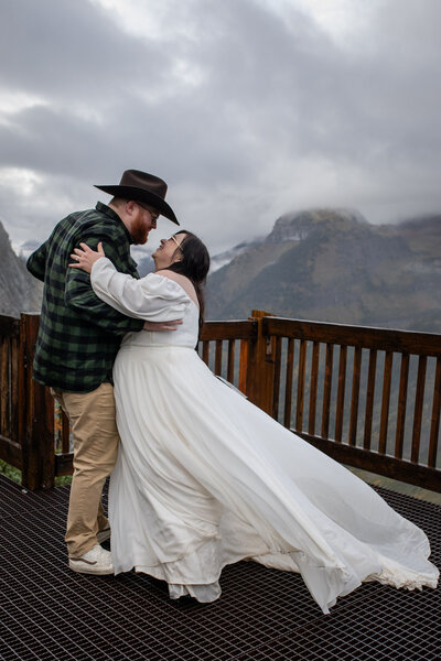 Newlyweds dance in the rain while standing on a wooden balcony with a massive valley below and peaks that disappear into the clouds.