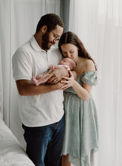 Mom in green dress kiss baby's head while dad in white and jeans is holding baby and smiling at him in studio in Annapolis Maryland photographed by Bethany Simms Photography