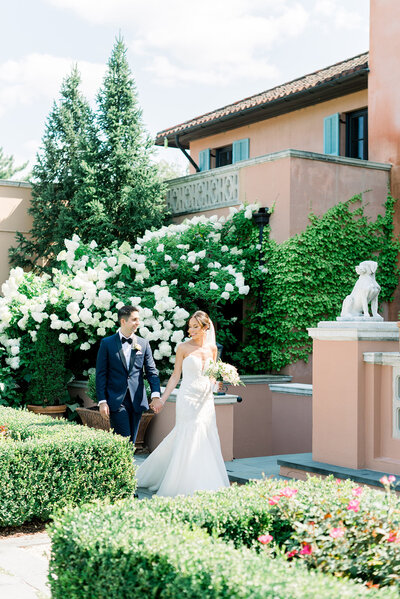 Newlyweds hold hands while walking through a flowering garden