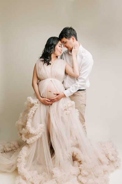 portrait of a husband and wife with their hands on her pregnant belly. They are in a studio wearing cream  with a white backdrop