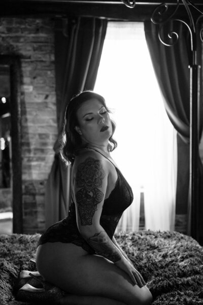 Black and white boudoir portrait of a woman in a black lace bodysuit kneeling on a bed