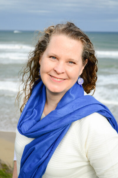 Sheila standing in front of the ocean wearing a blue  scarf smiling